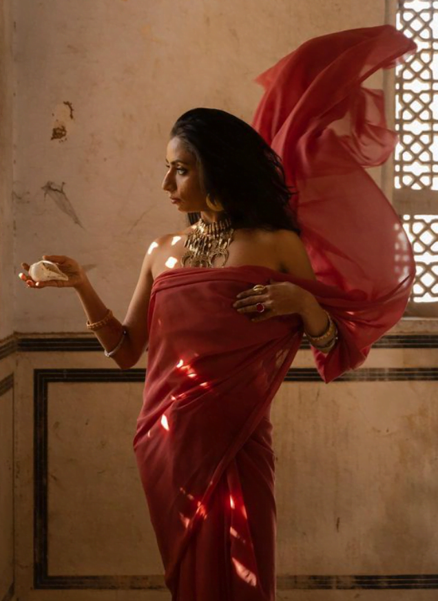 Shilo Shiv Suleman in a red saree standing holding a sea shell in one hand