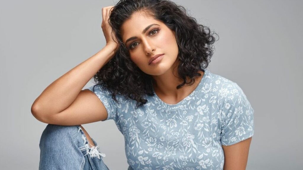 Kubbra Sait with her hand against her head posing for a photo in a blue top and jeans