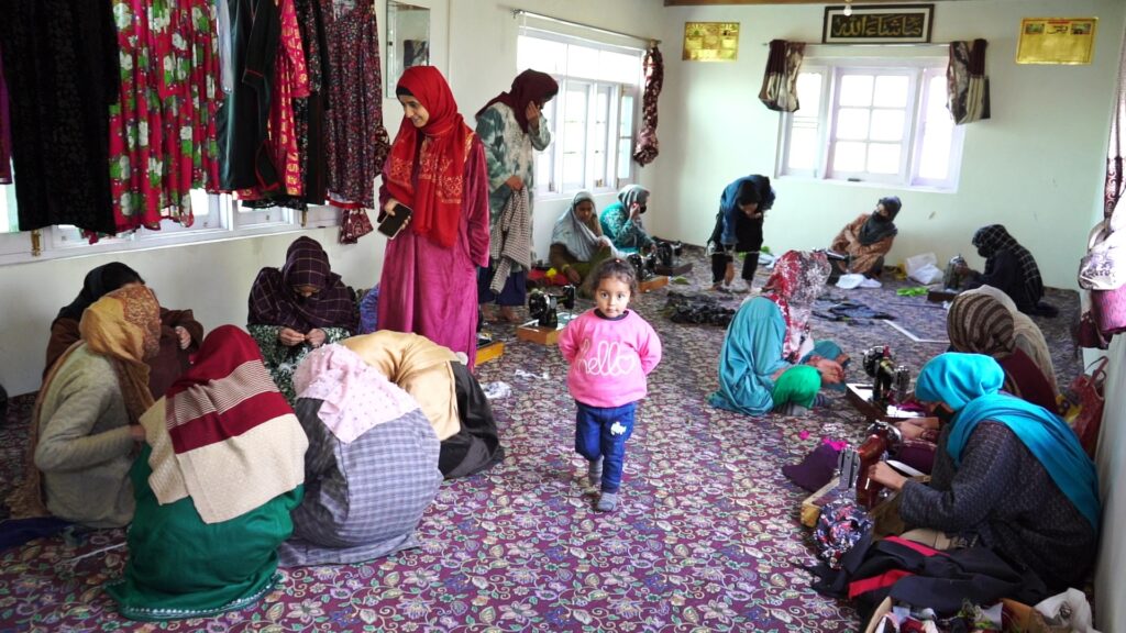 Gujjar women learning the art and craft of traditional Gujjar attire. A number of women sitting on floor with sweing machines in front. 